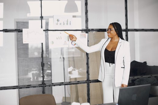 Woman in a white blazer pointing at printed bar and line charts taped to a glass office wall, laptop open on the desk beside her