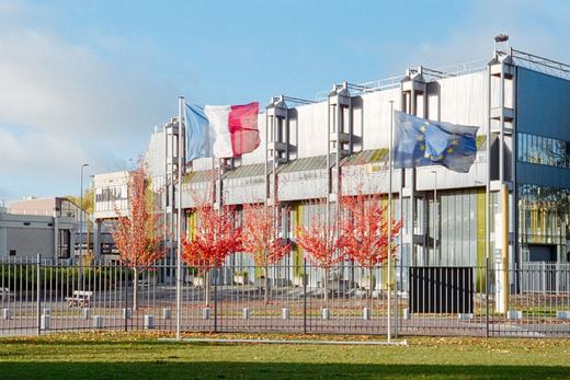 European Union and French flags flying in front of a modern glass-and-steel government building, autumn trees in the foreground