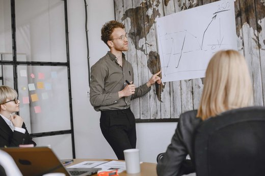 A regulatory professional presenting a hand-drawn trend chart on a flipchart to seated colleagues in front of a world-map wall mural