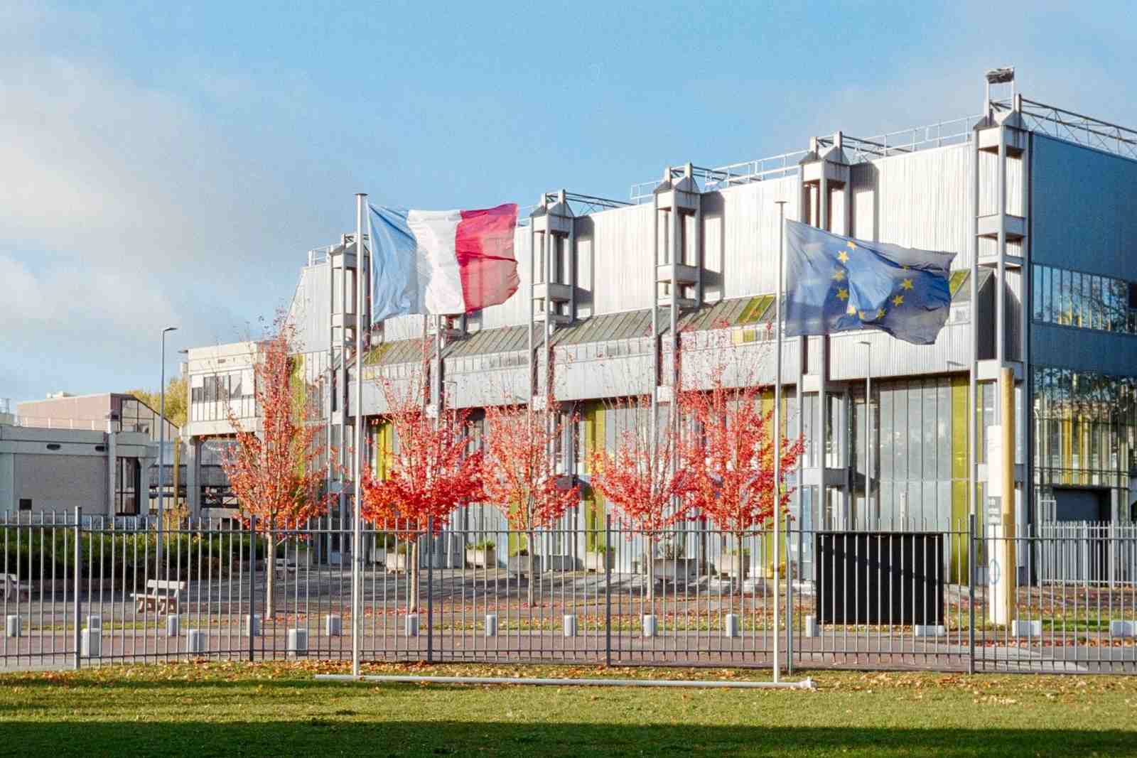 European Union and French flags flying in front of a modern glass-and-steel government building, autumn trees in the foreground