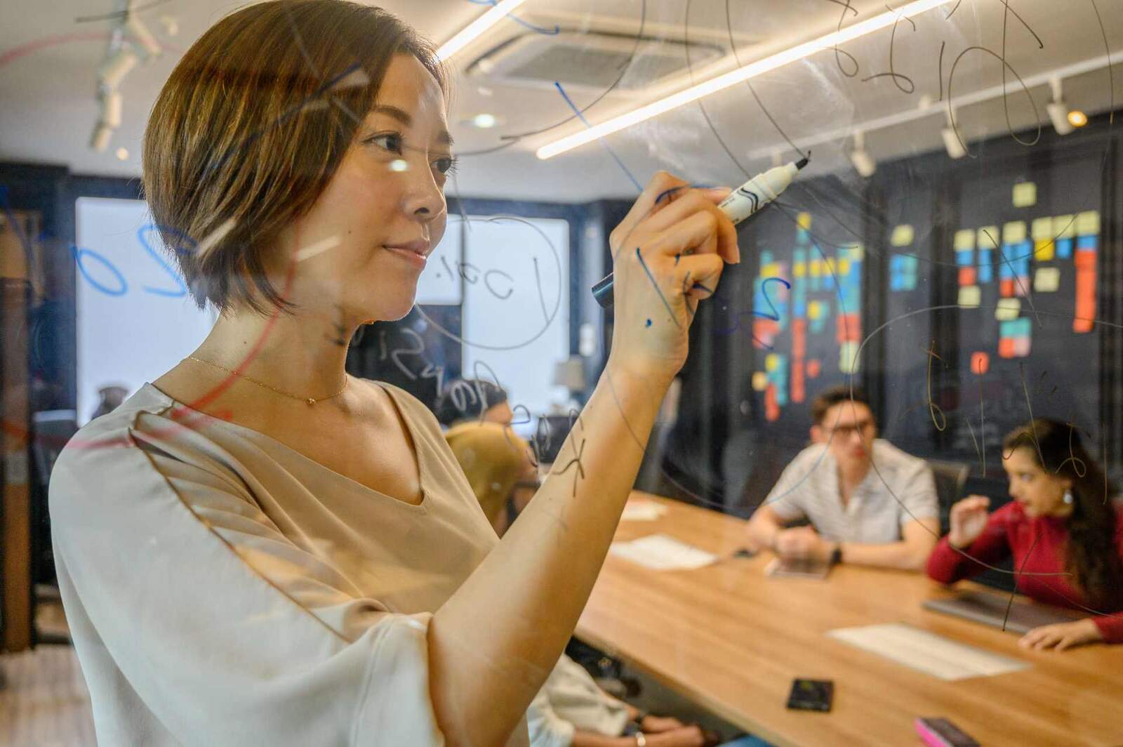 A regulatory professional writes on a glass wall with a marker during a team strategy session, colleagues reviewing documents at the table behind her