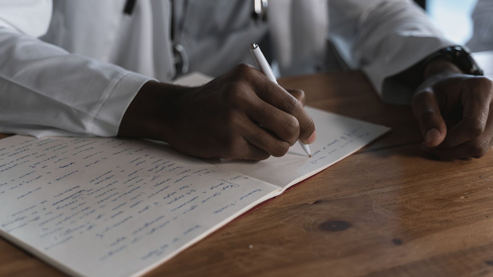 A physician in a white coat drafting notes by hand at a wooden desk