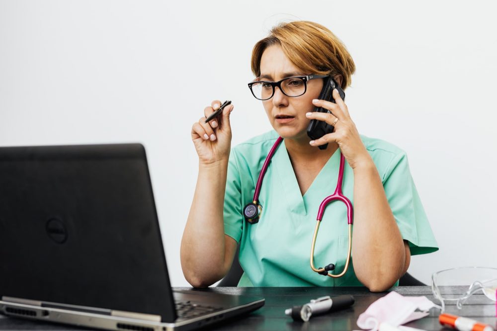 A clinician in scrubs working with a laptop and phone at a desk