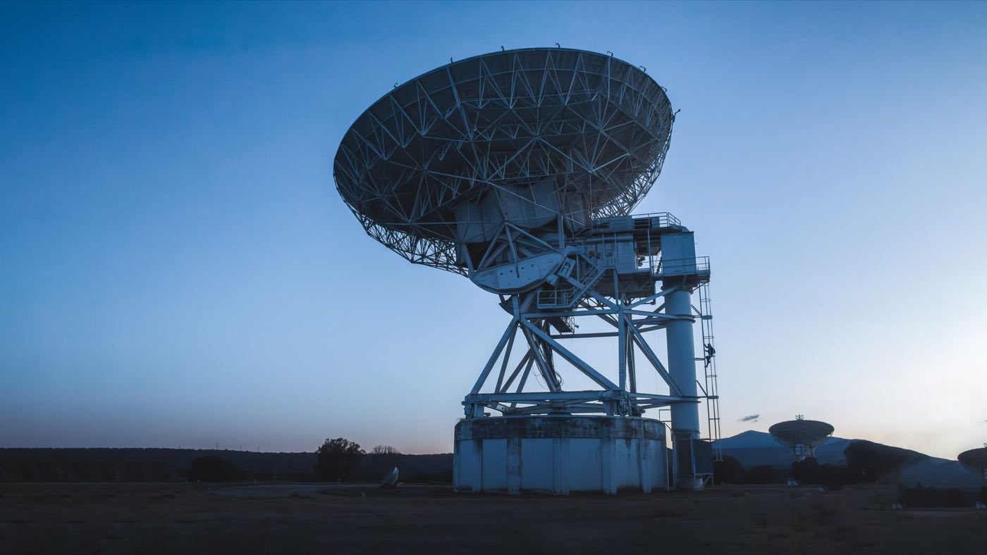 Ground-station antenna at dusk, transmitting like a regulatory submission gateway